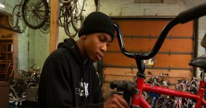 Man repairing a bicycle beside a recycle banner promoting environmental sustainability.