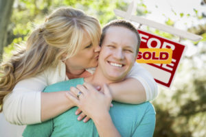 Happy Couple Hug In Front of Sold Real Estate Sign.